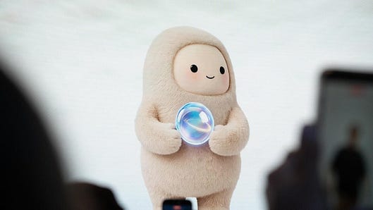 A soft, round digital assistant holds a glowing orb in a room with blurred people in the foreground.